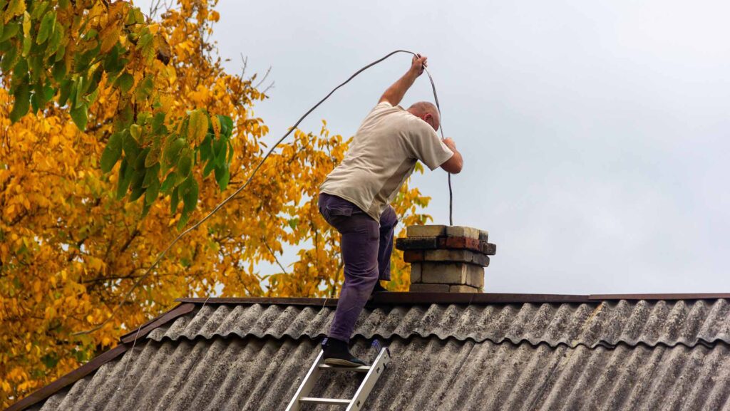 A man stands on a roof cleaning a chimney while standing on a ladder next to a tree with green and orange leaves.