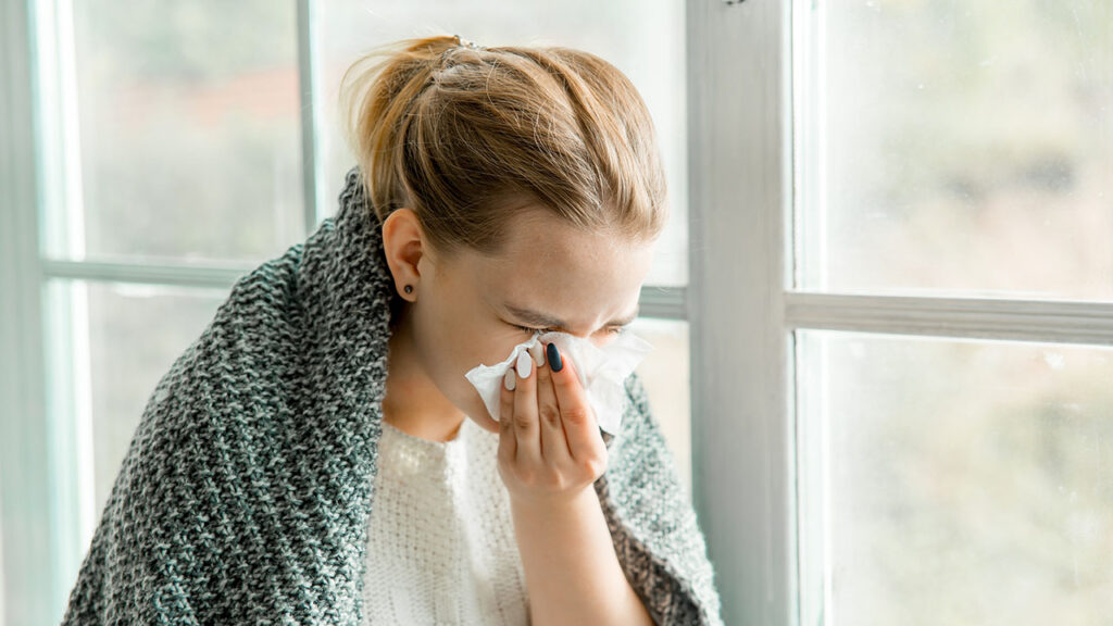 Woman wrapped in a blanket using a tissue to blow her nose.