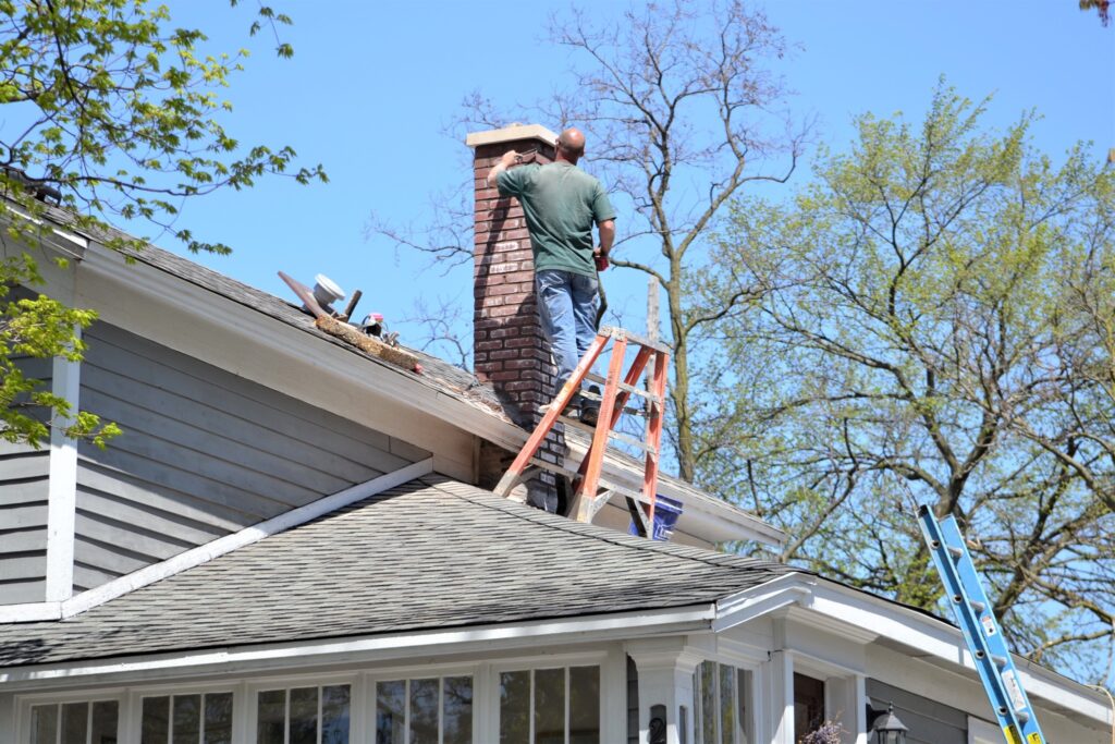 A man repairs a brick chimney while standing on the roof.