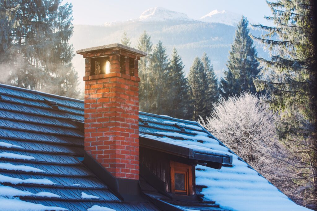 A brick chimney with smoke billowing out. The roof around it is snowy and there are mountains in the background.